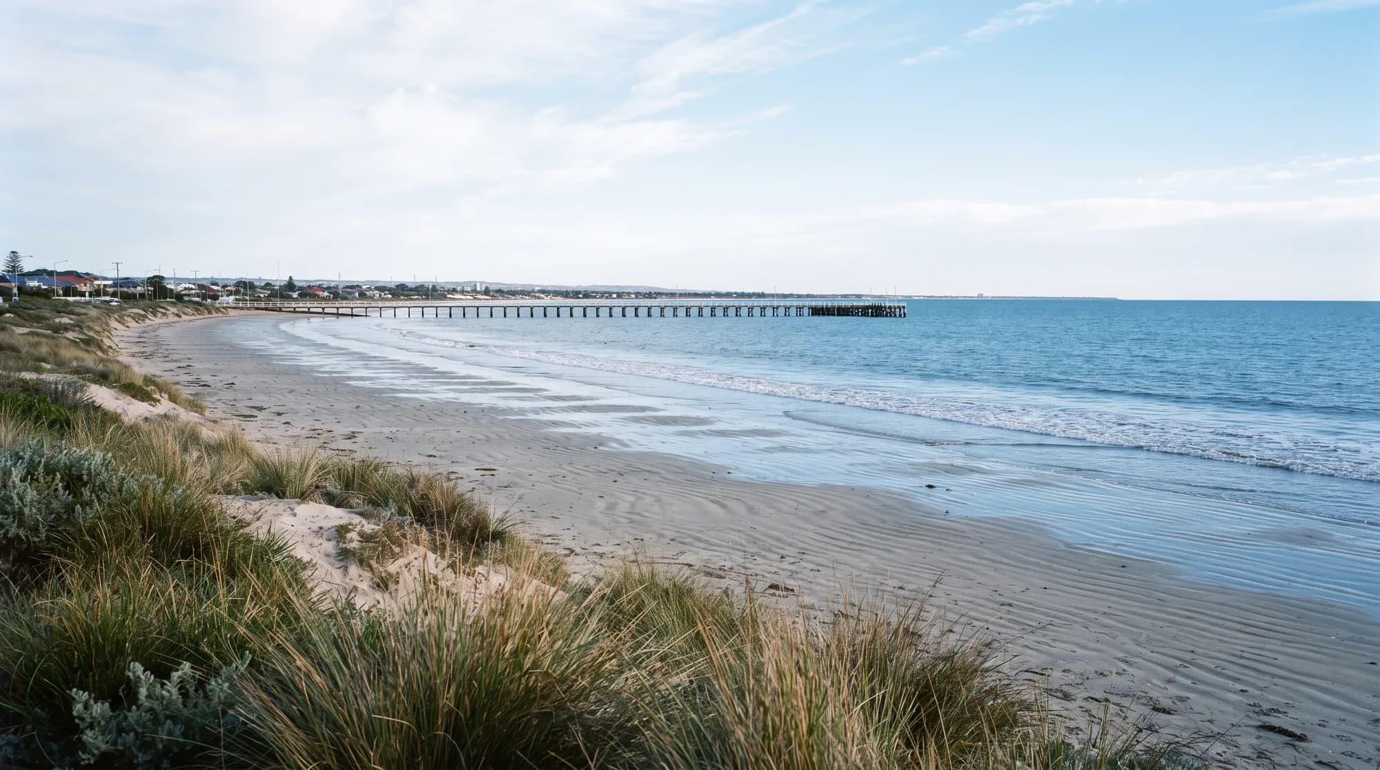 A wide sandy Adelaide beach under flat overcast light, with dune grass in the foreground and a long jetty running out to sea